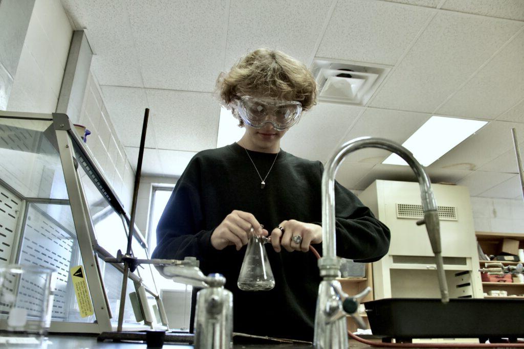 A person wearing safety goggles works with a glass flask in a laboratory setting, surrounded by various lab equipment and fixtures.