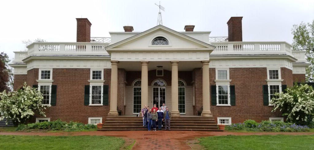 A group of people stand in front of a historic two-story brick mansion with white columns and green shutters under a cloudy sky.